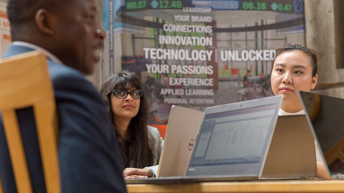 Two women at a table listen attentively during a business meeting with laptops open in front of them.