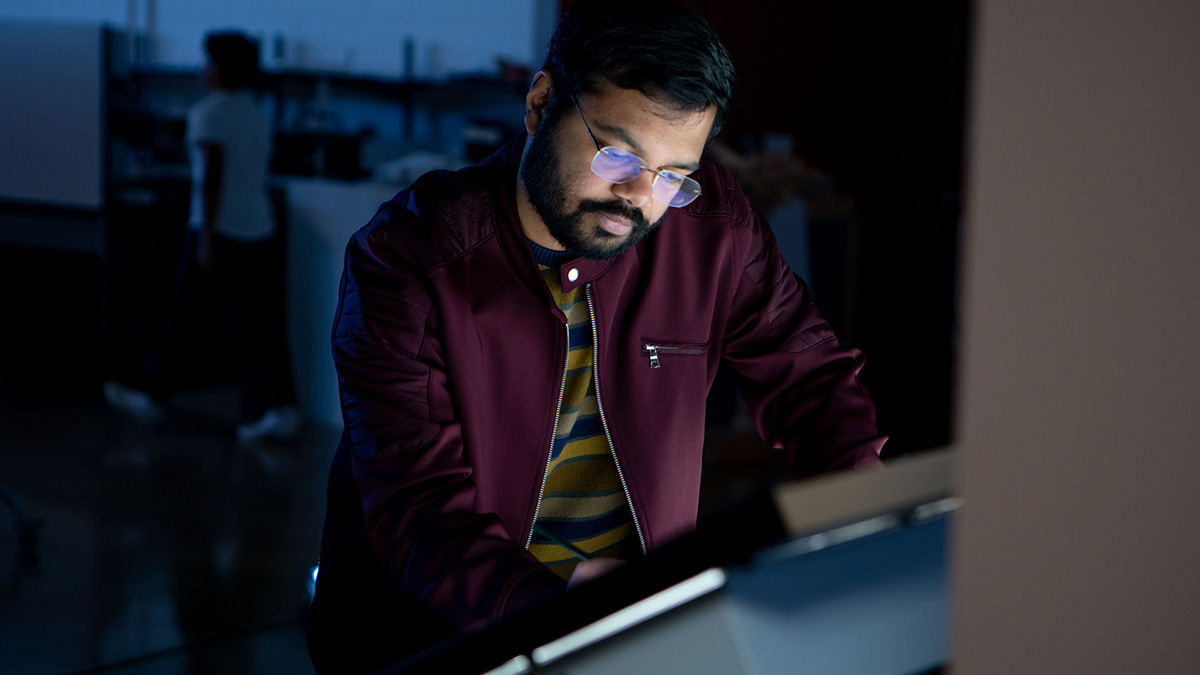 A person wearing glasses and a maroon jacket works intently at a digital touchscreen in a dimly lit room.