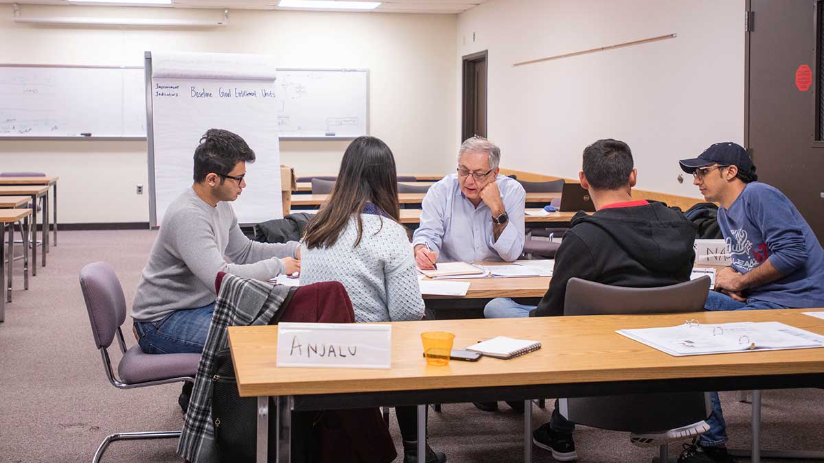 A small classroom group of students discusses coursework with an instructor seated around a table.