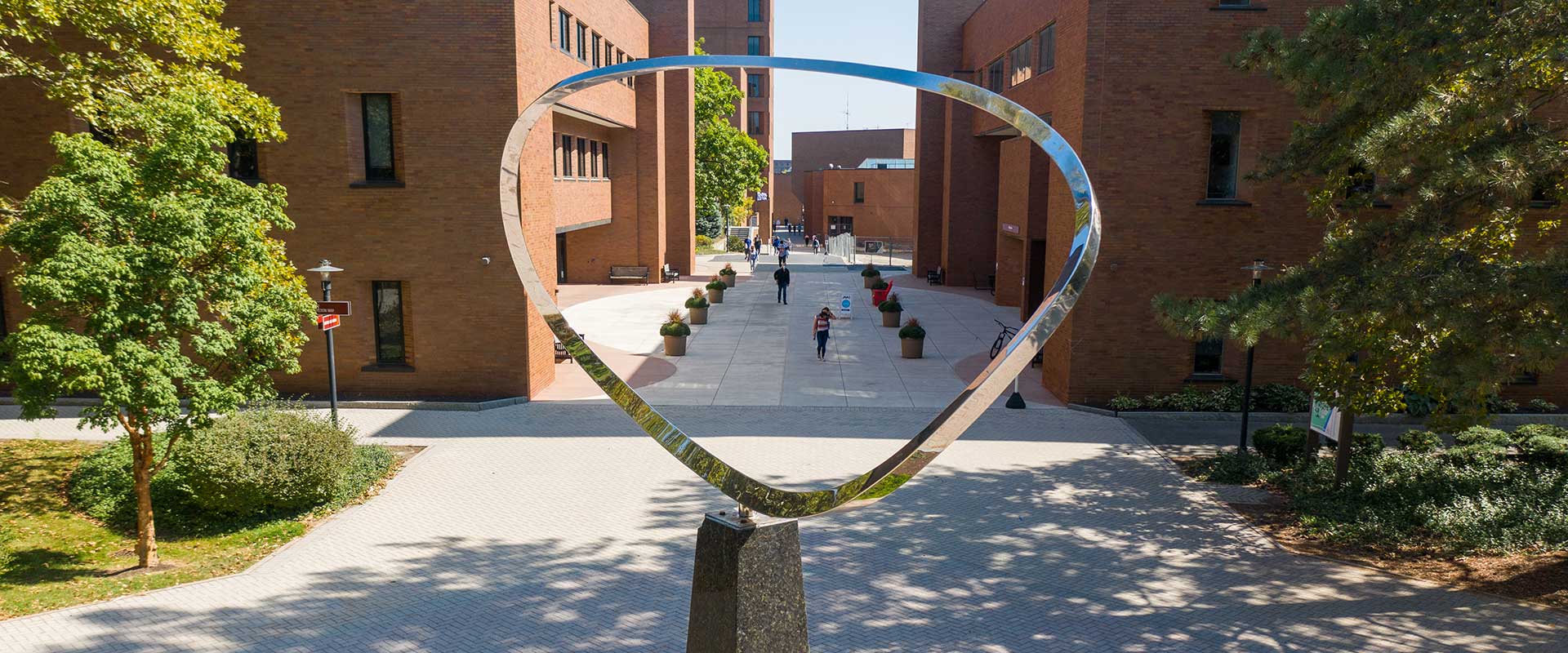 Pedestrians walk through a campus walkway framed by brick buildings and a large silver metal sculpture.