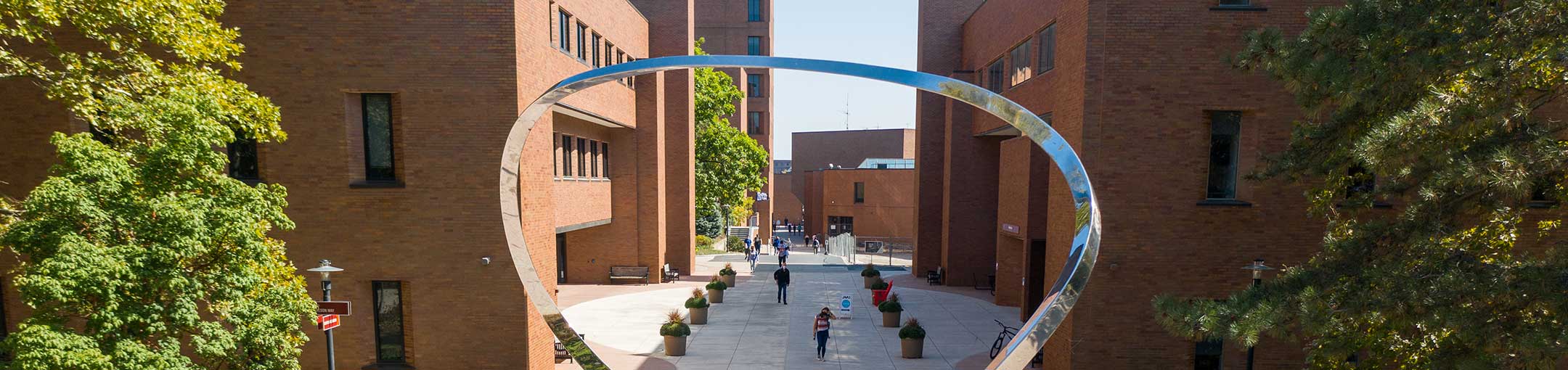 Pedestrians walk through a campus walkway framed by brick buildings and a large silver metal sculpture.