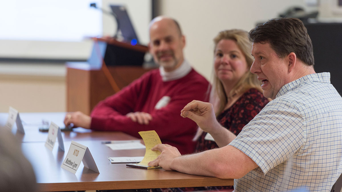 A discussion panel of participants in a classroom setting, one person speaking while others listen.