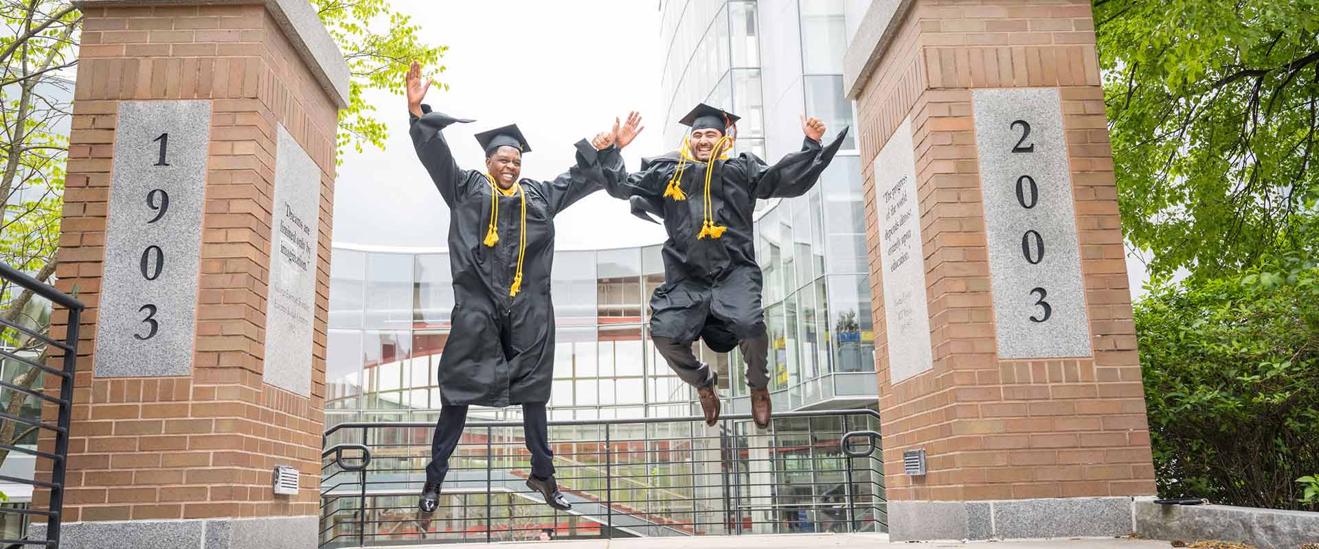 two recent grads wearing regalia jumping in the air in celebration.