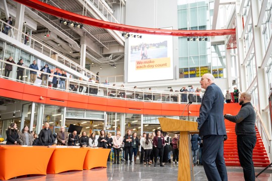 R I T president speaking at a podium in a large atrium with guests standing around.