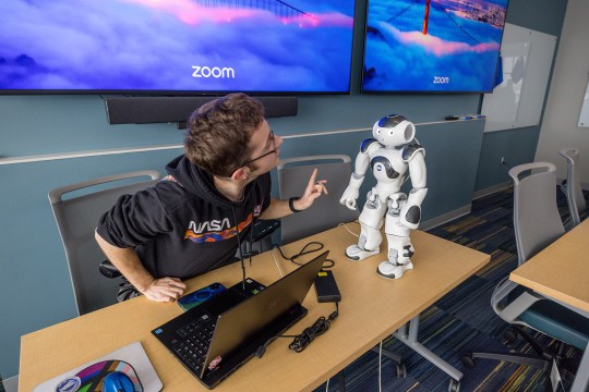 a student talks to a small robot on a table with computer screens behind him.