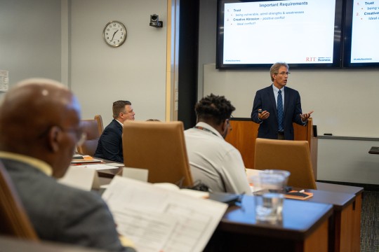 a man in a suit speaks to a room full of students with a projected display behind him.