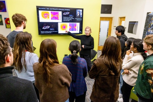 a woman with blonde hair points to a screen. in front of her, students gather to watch her speak.