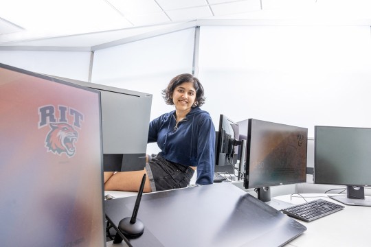 a woman sits on a desk surrounded by computer screens