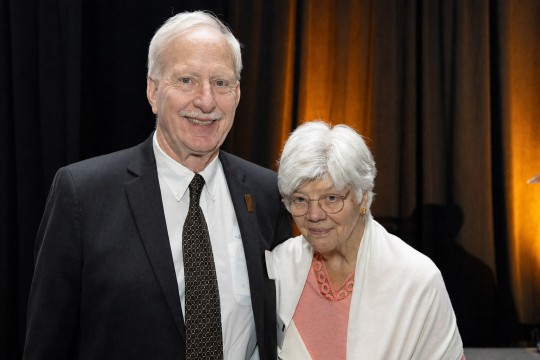 An older couple poses together at a formal event. The man wears a dark suit and patterned tie, while the woman wears a pink top with a white shawl. They stand in front of black drapes illuminated with warm amber lighting and smile toward the camera.