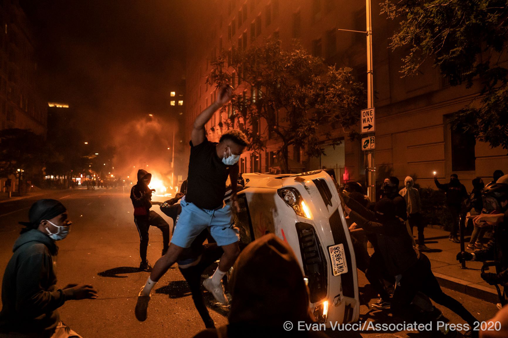 a photo from a riot where people are pushing a car over and there are fires in the background