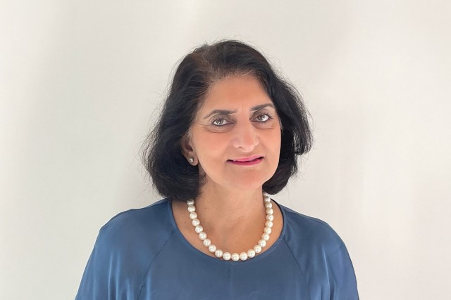 a dark haired woman in a blue shirt and pearl necklace stands in front of a white background.
