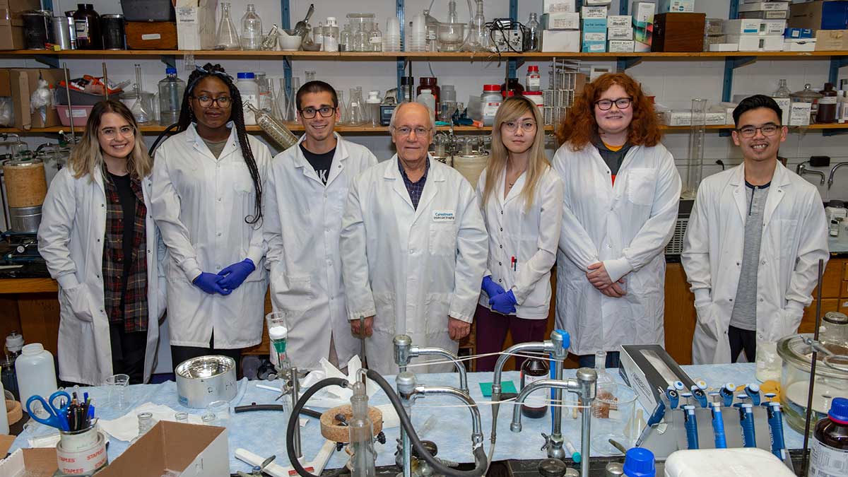 a professor and group of students wearing lab coats pose in a science lab.