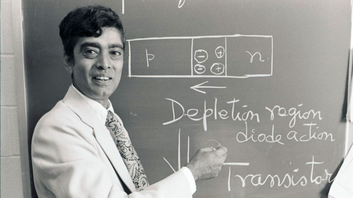 A man in a white suit points at a chalkboard diagram explaining transistor function in the 19 60s or 70s..