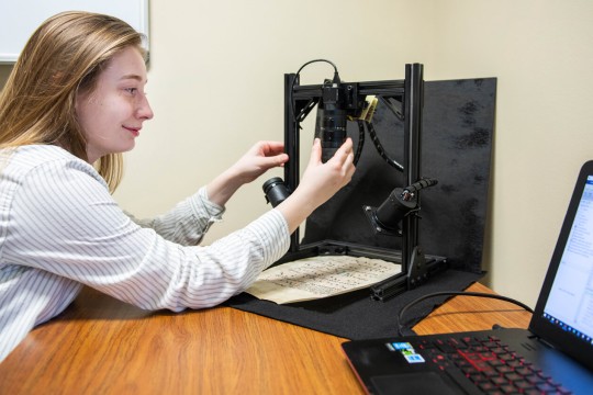 student adjusts cameras pointing to a manuscript next to a laptop.