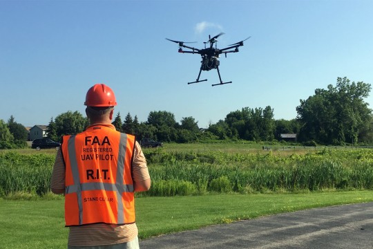 man wearing a vest identifying him as a U A V pilot flies a drone over a field.