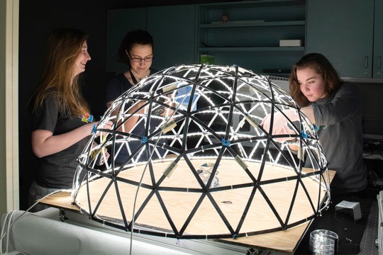 Three students stand around lighted spherical structure on a table.