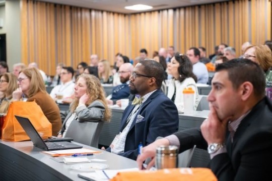 Dozens of business people sit in a lecture hall. One man wears a bowtie and suit jacket.