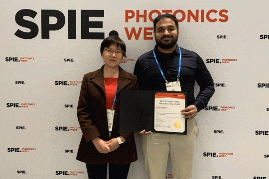 Two people stand in front of a banner, with man on right holding a certificate.