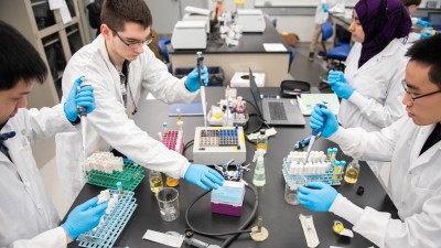 students in a lab setting wear lab coats use pipettes.