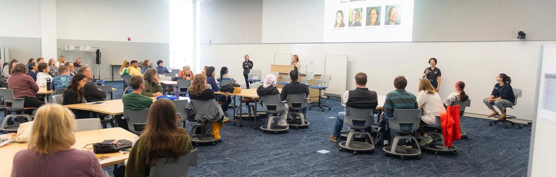 Students sitting around tables in a large classroom, watching a presentation.