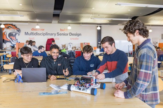 a group of students sit at a wooden table in a workshop, one works on a laptop while the others work on a robot car