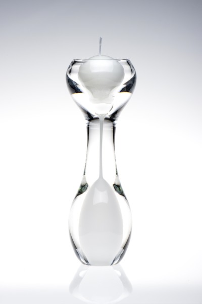 An hourglass shaped clear glass candle holder with white inside the bottom of the hourglass shape.