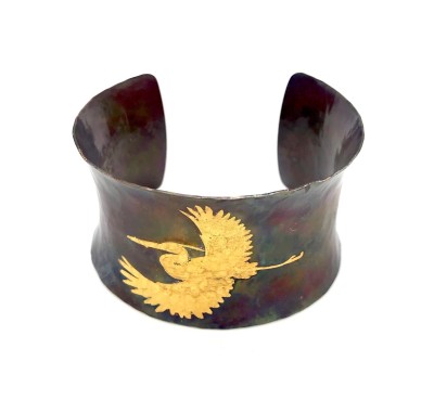 A wide copper cuff bracelet with a gold illustration of a heron in flight.