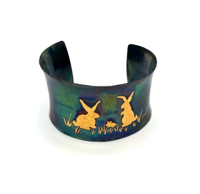 A wide copper cuff bracelet with a gold illustration of two bunny rabbits.