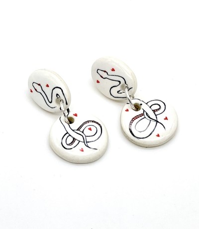 a pair of white ceramic dangle earrings with a round and oval disc and illustrations of a serpent surrounded by small red hearts.
