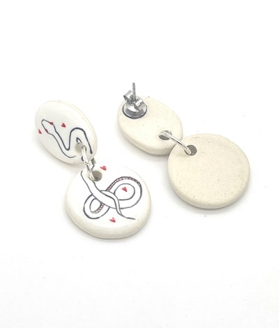a pair of white ceramic dangle earrings with a round and oval disc and illustrations of a serpent surrounded by small red hearts.