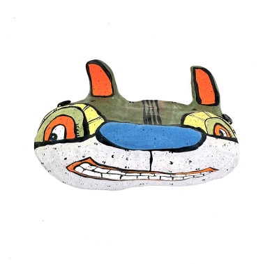 a hand sculpted ceramic mask form of a wide cat like face with orange ears, a blue nose, and a toothy smile.