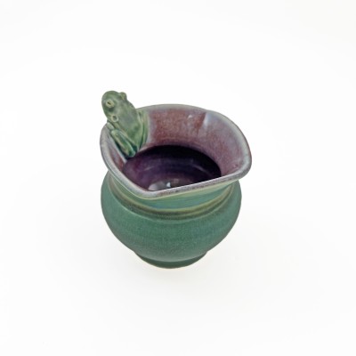 Turned view of small bud vase with square rim, burgundy interior, green exterior glaze, and a small frog perched on the rim
