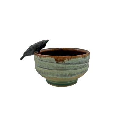 Side view of a small bowl with green hues and a brown interior, with a small black crow on the rim