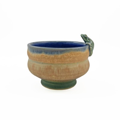 Side view of small bowl with a tan-orange exterior, with a deep blue interior glaze and a tiny frog perched on the rim