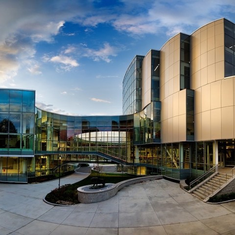 a panoramic view of a glass and steel clad modern building with a expansive walkway in front and puffy white clouds and blue skies above.
