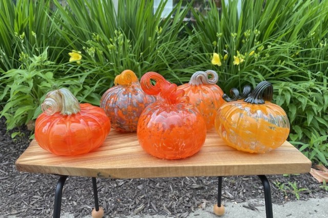 a group of five orange glass pumpkins on a wood platform in front of a green leafy background.