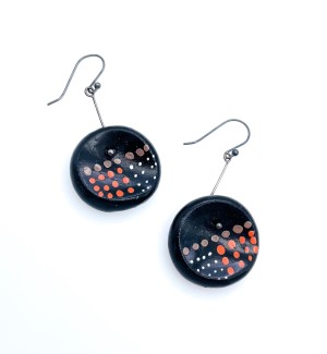 A pair of earrings with a black disk shaped clay part with red and brown dots hanging from a thin blackened silver wire.