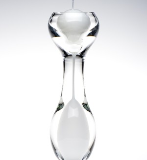 An hourglass shaped clear glass candle holder with white inside the bottom of the hourglass shape.