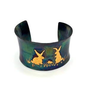 A wide copper cuff bracelet with a gold illustration of two bunny rabbits.