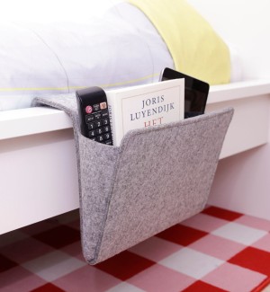 A grey felted pocket large enough to hold a laptop seen hanging off the side of a bed holding a book.