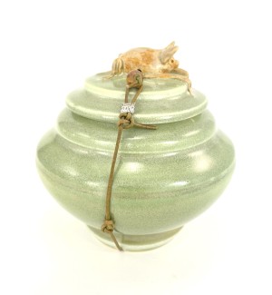 A green ceramic lidded jar with a small sculpture of a bat on top, tied with a waxed leather cord. 