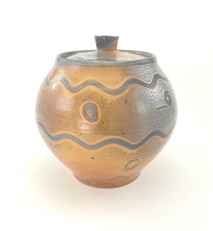 Rounded ceramic jar form with a lid and finial. Glazed is a burnt orange that meds into a medium grey. Surface design has wave pattern and circles around the circumference.