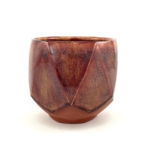 A low ceramic cup with an unglazed foot revealing the red clay body faceted surface. The upper portion is glazed in a rich mottled garnet colored glaze over the faceted surface.