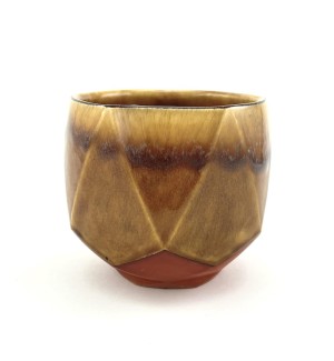 A low ceramic cup with an unglazed foot revealing the red clay body faceted surface. The upper portion is glazed in brown and ochre with charcoal toned highlights around the rim and along a few of the facet edges.