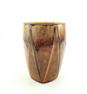 A tall ceramic tumbler with a faceted surface in brown and ochre glazes with charcoal toned highlights around the rim and along a few of the facet edges.