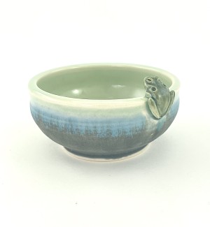 A low shallow ceramic bowl with a frog perched on the rim. The glaze blends from a moss green at the base to a ribbon of bright blue to celedon green on the lip and interior.
