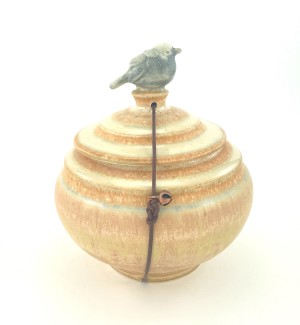 Round ceramic lidded peach glazed pot with sculpted bird on top. Lid is held with a leather cord, jasper stone and bell.
