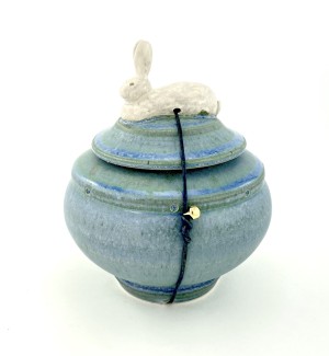 Rond lidded blue-green glazed ceramic pot with a rabbit form nestled top. Lid is secured with a leather cord, jasper stone and bell.