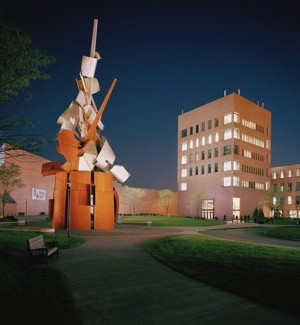 a color photograph of a large scale sculture of twisted metal on an open plaza with a brick building in the background with a midnight blue sky.