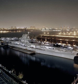 a full view of a large white battleship in a harbor with a nightime city skyline in the background.